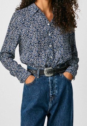 Kvinde iført en langærmet blå og hvid blomsterprint skjorte med knapper, stoppet ned i højtaljede blå jeans med et sort bælte med nitter.