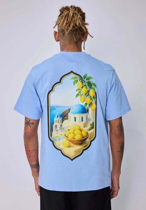 Personne portant une chemise bleu clair avec un motif d’une scène méditerranéenne montrant un bâtiment à coupole bleue, des citrons sur un arbre et dans un bol, ainsi que deux verres sur le rebord.