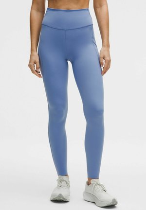 Kobieta ubrana w jasnoniebieskie legginsy z wysokim stanem oraz białe trampki, stojąca na jednolitym białym tle.