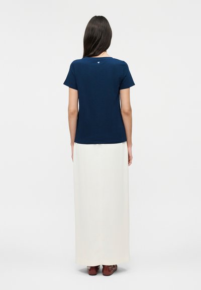 Γυναίκα που στέκεται με την πλάτη, φοράει μπλε σκούρο κοντομάνικο τοπ, μακριά λευκή φούστα και καφέ παπούτσια σε απλό φόντο.