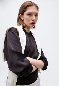 Jeune femme aux cheveux roux courts portant un chemisier noir et un pull blanc drapé sur les épaules, bras croisés, sur un fond clair uni.