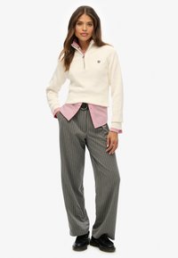 Frau steht mit Händen in den Taschen, trägt einen cremefarbenen Viertel-Reißverschluss-Pullover über einem rosa Shirt, graue gestreifte weite Hose und schwarze Schuhe.