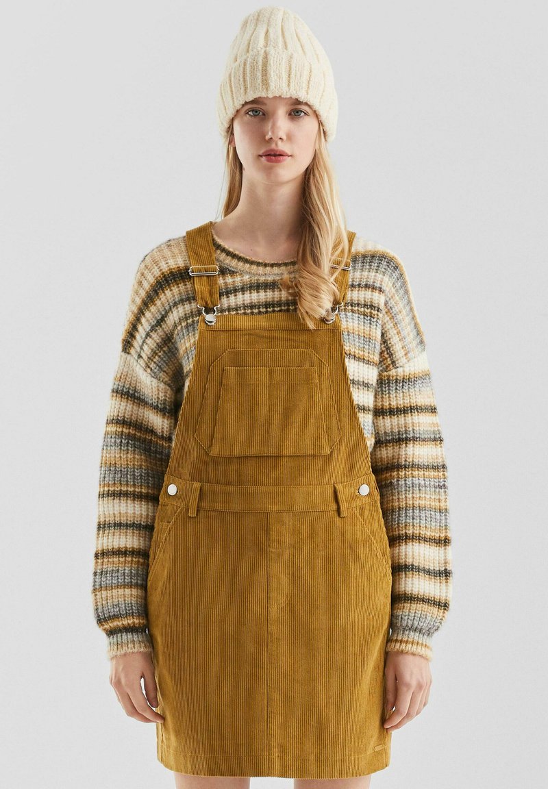 Mosterdgele corduroy overalljurk met een voorzak, gecombineerd met een gestreepte gebreide trui in beige, bruin en grijs tinten.
