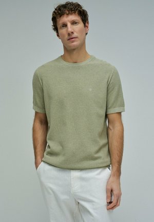 Joven con pelo rizado que lleva una camiseta de punto de manga corta de color verde claro y pantalones blancos, de pie con las manos en los bolsillos.