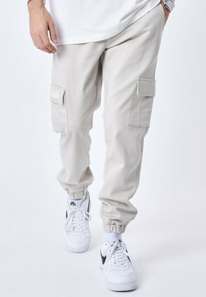 Pantalones cargo beige claro con puños elásticos, dos bolsillos laterales, parches de tela en contraste, combinados con zapatillas blancas.