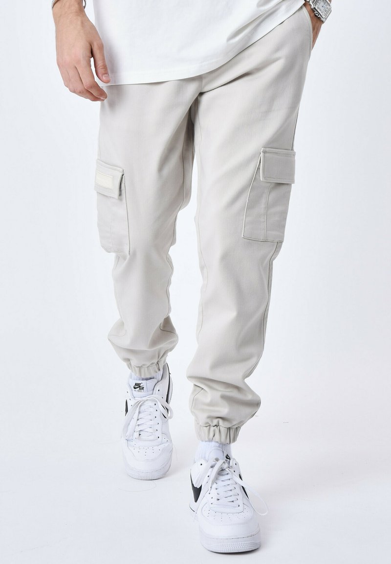 Pantaloni cargo beige chiaro con polsini elasticizzati, due tasche laterali, toppe in tessuto a contrasto, abbinati a sneakers bianche.