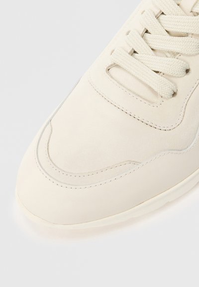 Close-up of a cream-colored sneaker showing the textured laces, stitched suede upper, and smooth sole edge against a light background.
