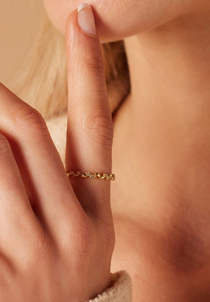 Gouden ring met kleine heldere accenten op een slanke vinger, tegen een neutrale achtergrond. Huidstructuur zichtbaar op de hand.