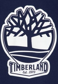 Logo Timberland su un tessuto blu navy. Presenta una silhouette bianca di un albero con rami geometrici e testo sottostante: "Timberland Est. 1973."