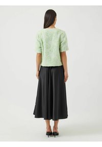 Lys grønn teksturert kortermet jakke med blomsterdesign, sammen med et langt svart skjørt, sett fra bak. Svarte hæler synlige.