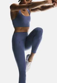 Kvinde i blå sportstøj strækker armene frem, mens hun løfter det ene knæ, iført hvide atletiske sko på en hvid baggrund.