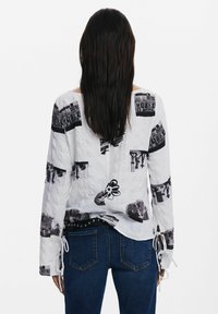 Cămașă albă cu mâneci lungi, cu imprimeuri grafice negre și textură cu caneluri, prezentând șnururi ajustabile la mâneci și un croi lejer.