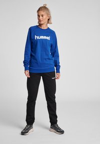 Felpa blu con logo bianco "hummel", abbinata a pantaloni della tuta neri con un piccolo logo. Il modello indossa sneaker nere e grigie.