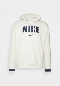 Krämfärgad huvtröja med en kenguruficka, oversized passform, marinblå "NIKE"-logotyp framtill och marinblå manschettaccent på ärmarna. Mjuk tygtextur.
