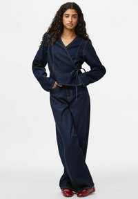 Sieviete ar cirtainiem matiem, tērpusies tumši zilā wrap tipa topā un platajam džinsiem, stāv ar vienu roku kabatā un valkā sarkanas kurpes.