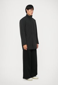 Sort frakke med høj krave og lynlås foran, med sidelommer. Accepteret af løse sorte bukser og off-white sko, mod en ensfarvet baggrund.