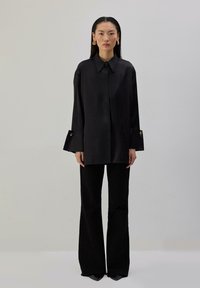 Γυναίκα με μακριά σκούρα μαλλιά που φοράει υπερμεγέθη μαύρο πουκάμισο με μανσέτες με χρυσά κουμπιά, μαύρο παντελόνι φαρδιάς γραμμής και χρυσά σκουλαρίκια, στέκεται μπροστά σε απλό τοίχο.