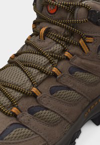 Καφέ ορειβατικό μποτάκι με κορδόνια σε μαύρο και κίτρινο σχέδιο, πάνελ από υφαντό ύφασμα και πορτοκαλί λογότυπο Merrell στην πλευρά.