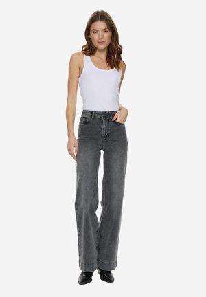 Femeie purtând un top alb fără mâneci, blugi negri decolorați, cu talie înaltă, și pantofi negri cu vârf ascuțit, stând cu o mână în buzunar.