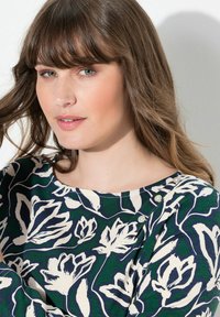 Žena s dlouhými hnědými vlasy a ofinou, oblečená v zeleném a námořnickém květinovém vzoru s perlovými knoflíky u výstřihu.