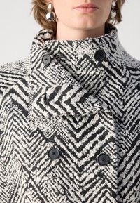Abrigo texturizado en blanco y negro con patrón de chevrones, que cuenta con un alto cuello y dobles botones negros. Tejido suave, de textura entrelazada y superficie esponjosa.