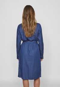 Jeans obleka s dolgimi rokavi, pasom in dolžino do kolen v enotni temno modri barvi. Gladka tekstura, zakopčane manšete.