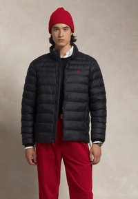 Zwarte pufferjack met horizontale padding, ritssluiting en klein rood logo, gecombineerd met een witte shirt en rode broek. Rode beanie op het hoofd.