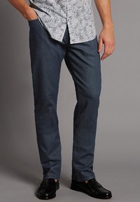 Pantalones de mezclilla azul marino con un ajuste slim, que cuentan con bolsillos y una textura suave, combinados con zapatos negros pulidos y una camisa de estampado.