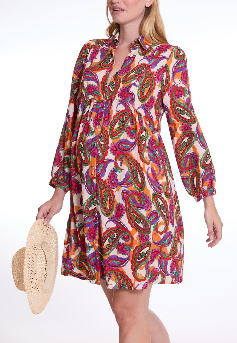 Bunt gemustertes Kleid mit Paisley-Muster, langen Ärmeln, kragenartigem Hals und lockerer Passform. Eine Frau hält einen gewebten Strohhut.