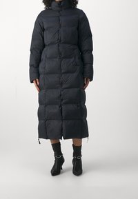 Dlouhý černý péřový kabát s prošívanými panely, vysokým límcem a předními knoflíky; boční zipy; nošený s černými kotníkovými botami.