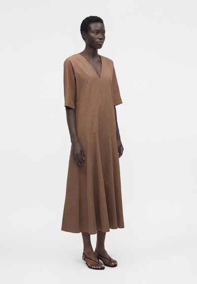 Γυναίκα με κοντά σγουρά μαλλιά φοράει ένα άνετο καφέ φόρεμα μέχρι τη μέση της γάμπας με μανίκια μέχρι τον αγκώνα και μαύρα σανδάλια με τακούνι, στέκεται μπροστά σε λευκό φόντο.