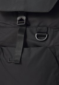 Zwarte rugzak met een zachte nylon buitenkant, een bovenflap sluiting met een plastic gesp, geweven schouderband en een metalen D-ring accent.