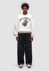 Felpa bianca con stampa e design grafico "WRSTBHVR", abbinata a pantaloni neri larghi e sneakers multicolori. Colori solidi, vestibilità comoda.