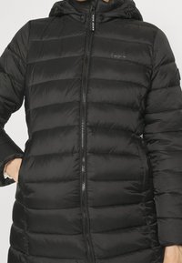 Abrigo acolchado negro con cierre frontal, capucha y bolsillos laterales. Presenta una textura acolchada y una marca en el pecho.