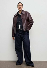 Bordowa skórzana kurtka typu cropped z szerokim kołnierzem, zestawiona z ciemną denimową koszulą i luźnymi, ciemnoniebieskimi dżinsami. Klasyczne guziki.