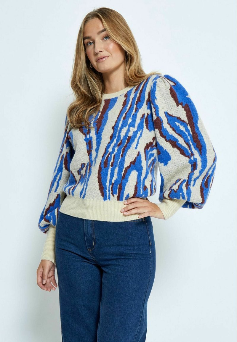 Strickpullover mit cremiger Basis, der blaue und braune abstrakte Muster sowie puffärmliche Ärmel aufweist. Kombiniert mit dunkelblauen Jeans.