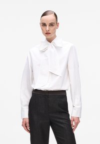 White long-sleeve blouse with a large bow tie collar, paired with dark textured trousers featuring a smooth waistband. Minimalist design.
