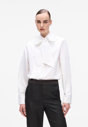 White long-sleeve blouse with a large bow tie collar, paired with dark textured trousers featuring a smooth waistband. Minimalist design.