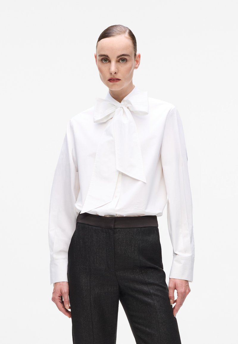 White long-sleeve blouse with a large bow tie collar, paired with dark textured trousers featuring a smooth waistband. Minimalist design.