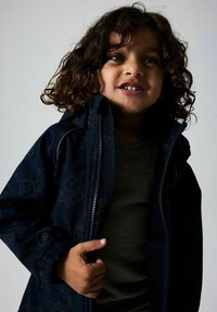 Veste marine à motif avec capuche et fermeture éclair sur le devant, fabriquée en matériau léger. Chemise intérieure grise visible avec texture côtelée.
