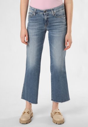 Vrouw met lichtblauwe cropped flare jeans, beige loafers en een roze top, staand tegen een effen witte achtergrond.