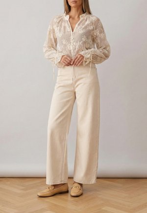 Kvinde iført gennemsigtig beige bluse med blomsterbroderi, cremefarvede vidde bukser og brune loafers, stående på trægulv.
