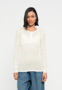 Κρεμ τοπ με μακριά μανίκια, ραβδωτή υφή, σχεδιασμό σε σχήμα V και λαιμόκοψη με κουμπιά, συνδυασμένο με άνετο μπλε τζιν παντελόνι.