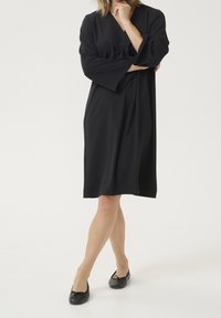 Robe noire en tissu lisse, avec un décolleté en V, des manches larges et une taille élastique. Associée à des ballerines noires.