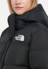 Crna puffer jakna s visokim ovratnikom, sa bijelim logotipom The North Face. Tkanina je glatka s podstavljenom teksturom.