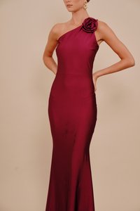 Bordo krāsas kleita ar vienu plecu, izgatavota no gluda auduma, ar rožu detaļu pie pleca un pieguļošu siluetu, kas paplašinās pie apakšas.