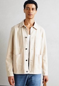 Veste beige clair en coton avec un col, fermeture à boutons, deux poches avant et une coupe décontractée, associée à un jean bleu.