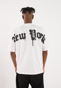 Camiseta blanca de gran tamaño con gran texto gótico negro "New York" en la espalda, mangas cortas y una suave textura de algodón.