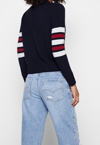 Pull en maille marine avec des accents à rayures blanches et rouges sur les manches. Assorti à un jean en denim bleu clair usé, affichant une coupe décontractée.