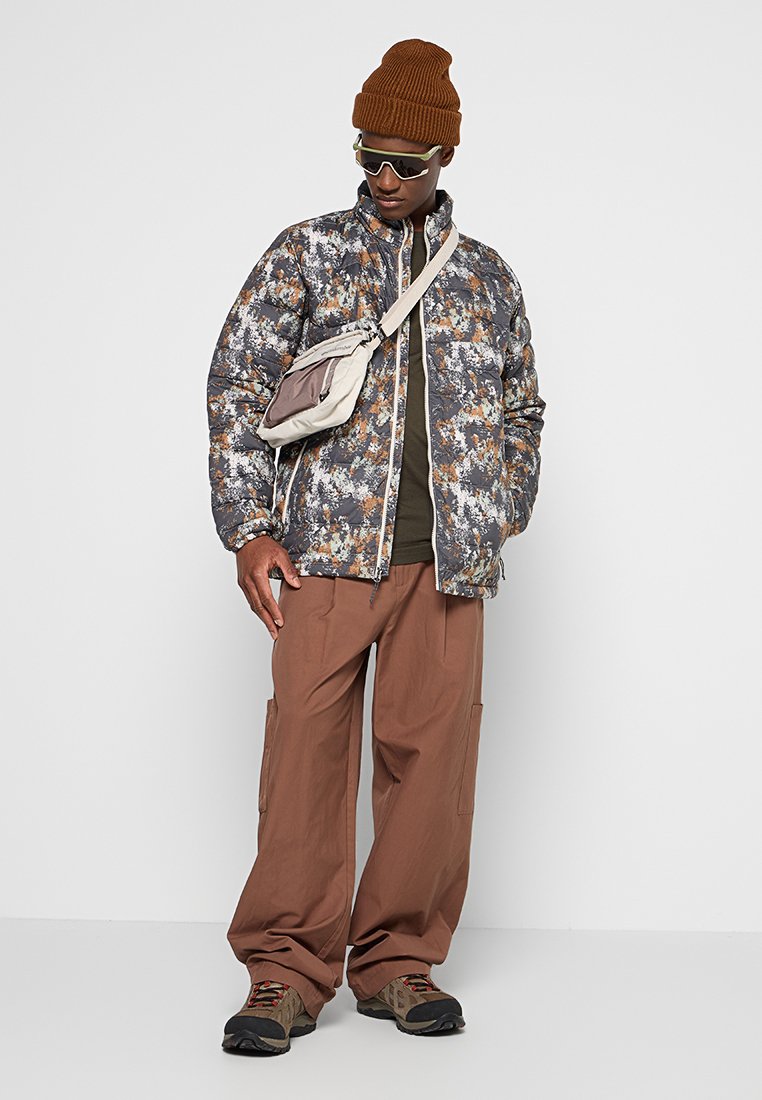 Kurtka puchowa w szarym, brązowym i pomarańczowym wzorze kamuflażu; brązowe spodnie cargo; beżowa torba na ramię; dzianinowa brązowa czapka; ciemne okulary przeciwsłoneczne.